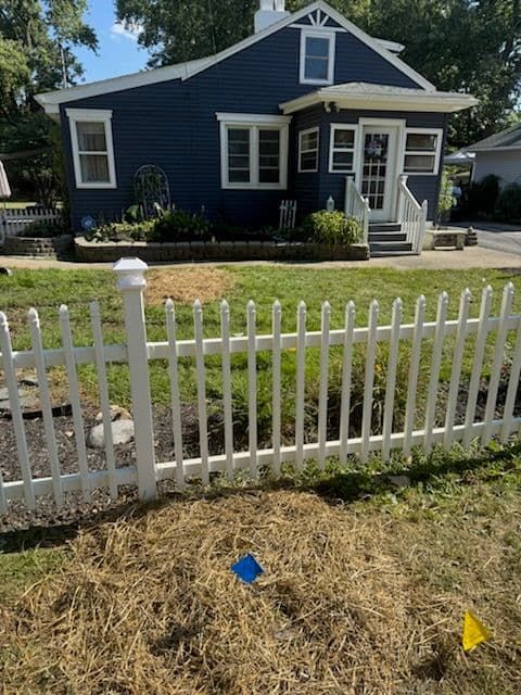 Blue house with white picket fence, grassy lawn, and blue and yellow marker flags.