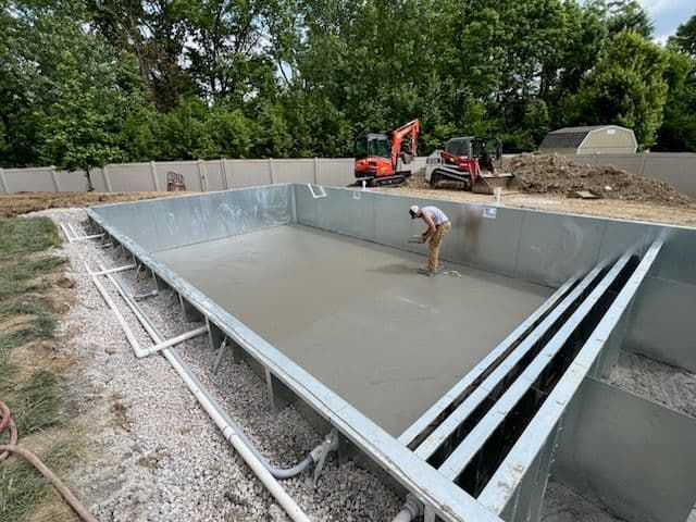 Man smoothing cement in a metal-framed pool under construction; two excavators in the background.