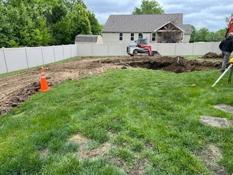 Backyard construction site with a skid steer digging; a fence surrounds the area.