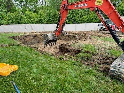 A Kubota excavator digs a hole in a grassy yard, preparing for construction, near a white fence.
