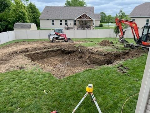 Construction site with backhoe and skid steer digging in backyard, white fence and house in background.