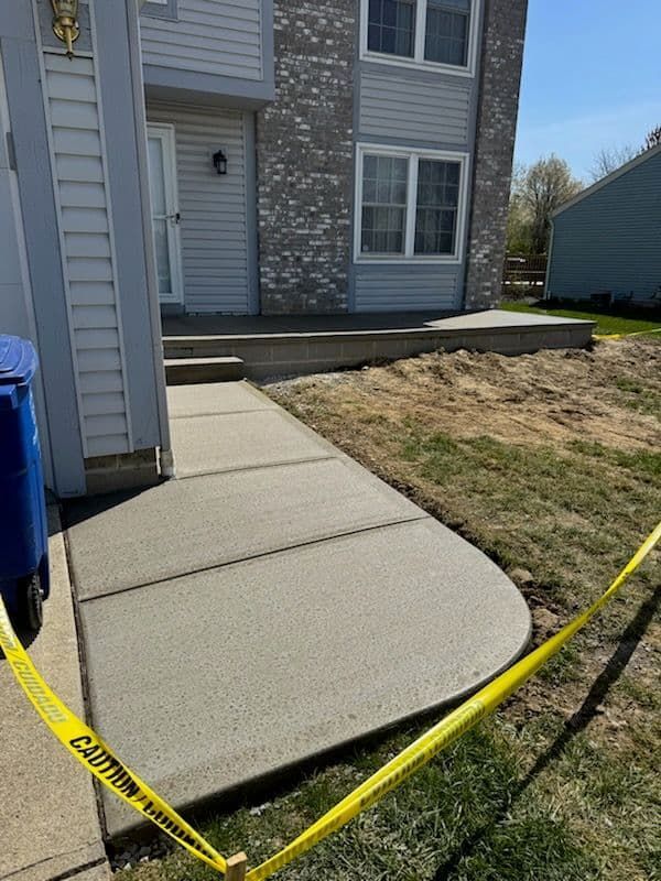 Newly poured concrete walkway leading to a two-story house. Yellow caution tape surrounds the walkway.
