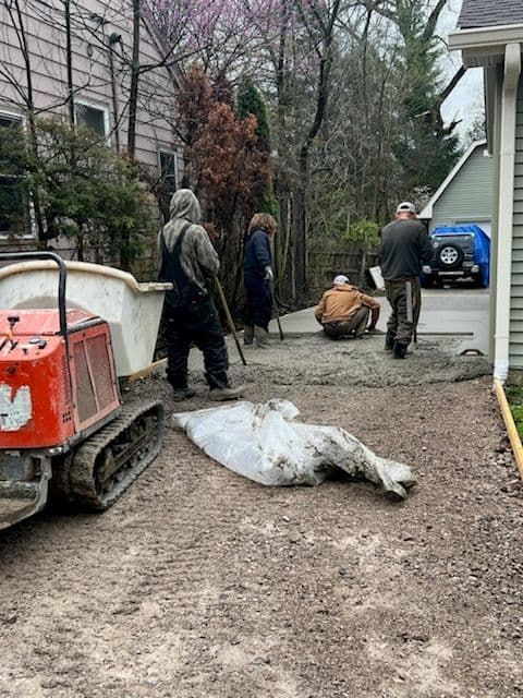 Construction crew working on a driveway. One operating mini-excavator, others preparing concrete. Outdoors, cloudy day.