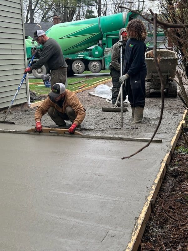 Construction workers pouring and leveling concrete for a patio next to a house; a concrete truck in background.