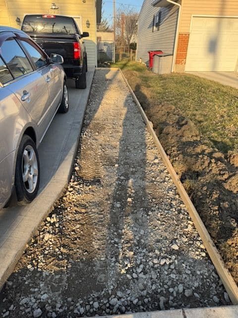 A narrow driveway under construction with concrete borders, soil and gravel filling it. Two cars parked nearby.