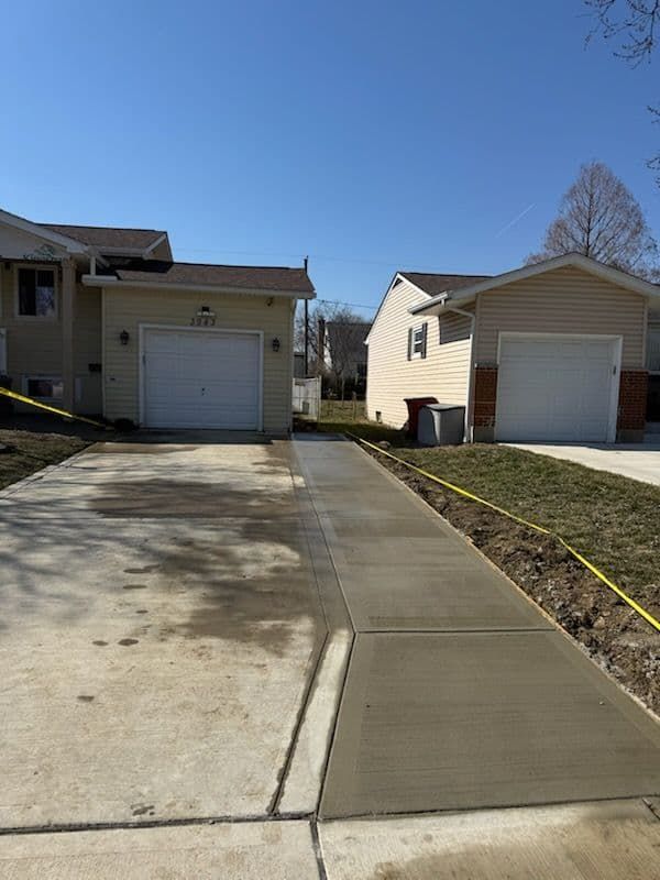 New concrete driveway and sidewalk between two houses with attached garages, bright blue sky.