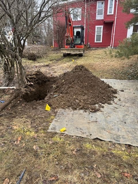 Excavator digging in a yard, next to a pile of dirt and a red building.