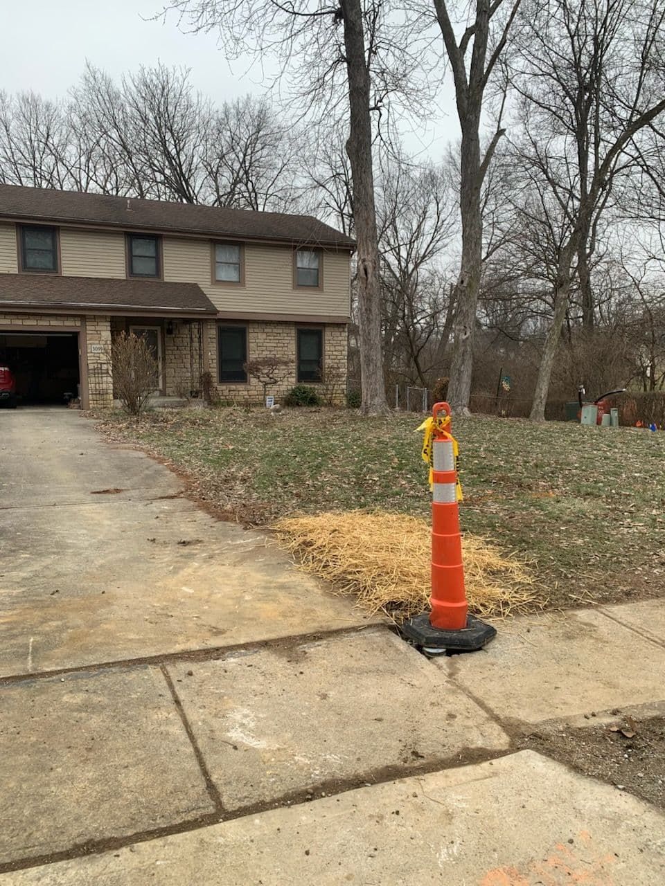 Two-story house with yard. Orange cone on sidewalk; grass and bare trees in front. Cloudy day.