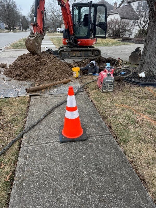 Construction site with excavator, orange cone, worker in a hole, and equipment on a sidewalk.