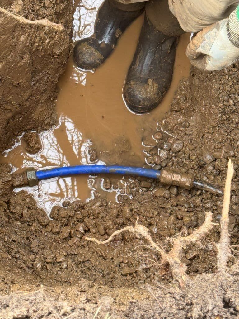 Person in muddy boots in a ditch inspecting a blue pipe connection in dirt.