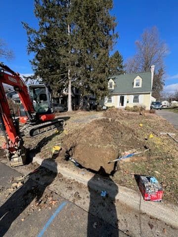 A small excavator digs in a front yard, with a pile of dirt, and a house in the background.