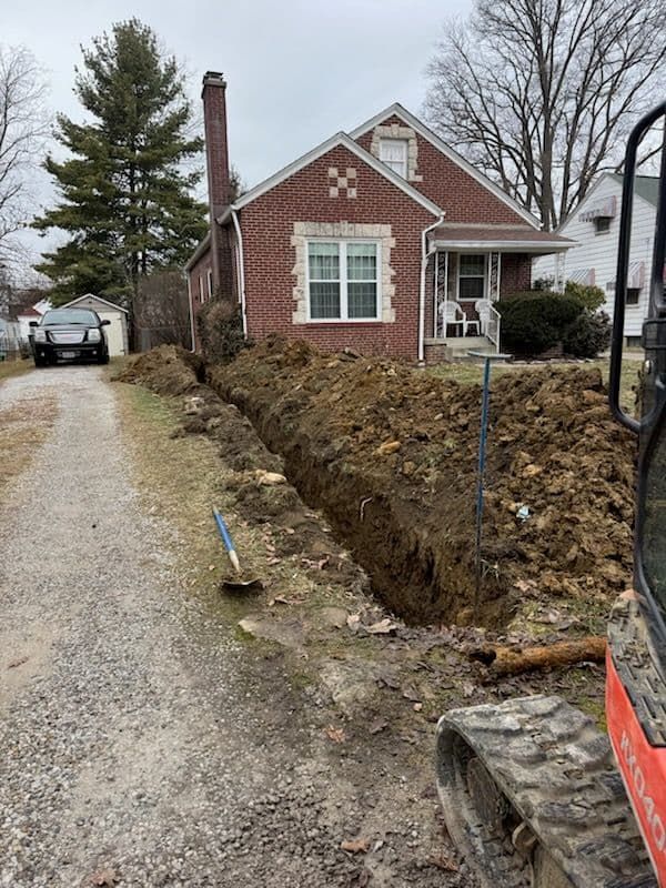 A trench dug along the driveway of a brick house; a small excavator is visible in the right corner.