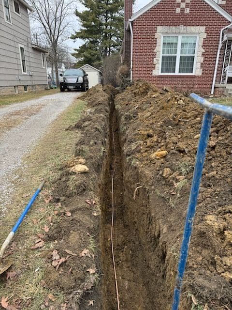 Trench dug alongside a driveway, leading to a red brick house. Blue-handled tools rest nearby.