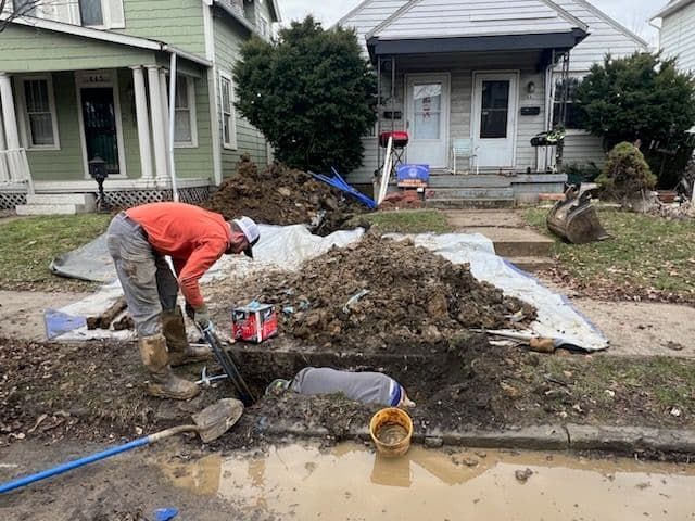 Two workers are repairing a pipe in a residential yard, surrounded by dirt and tools.
