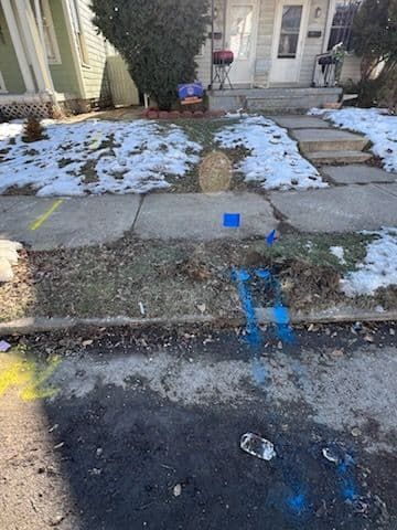 Sidewalk with utility markings; snow-covered yard in front of a house. Yellow and blue paint lines indicate underground utilities.