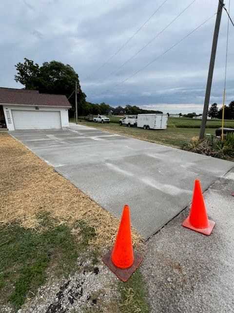 Newly poured concrete driveway in front of a garage, with orange safety cones. Cloudy day.
