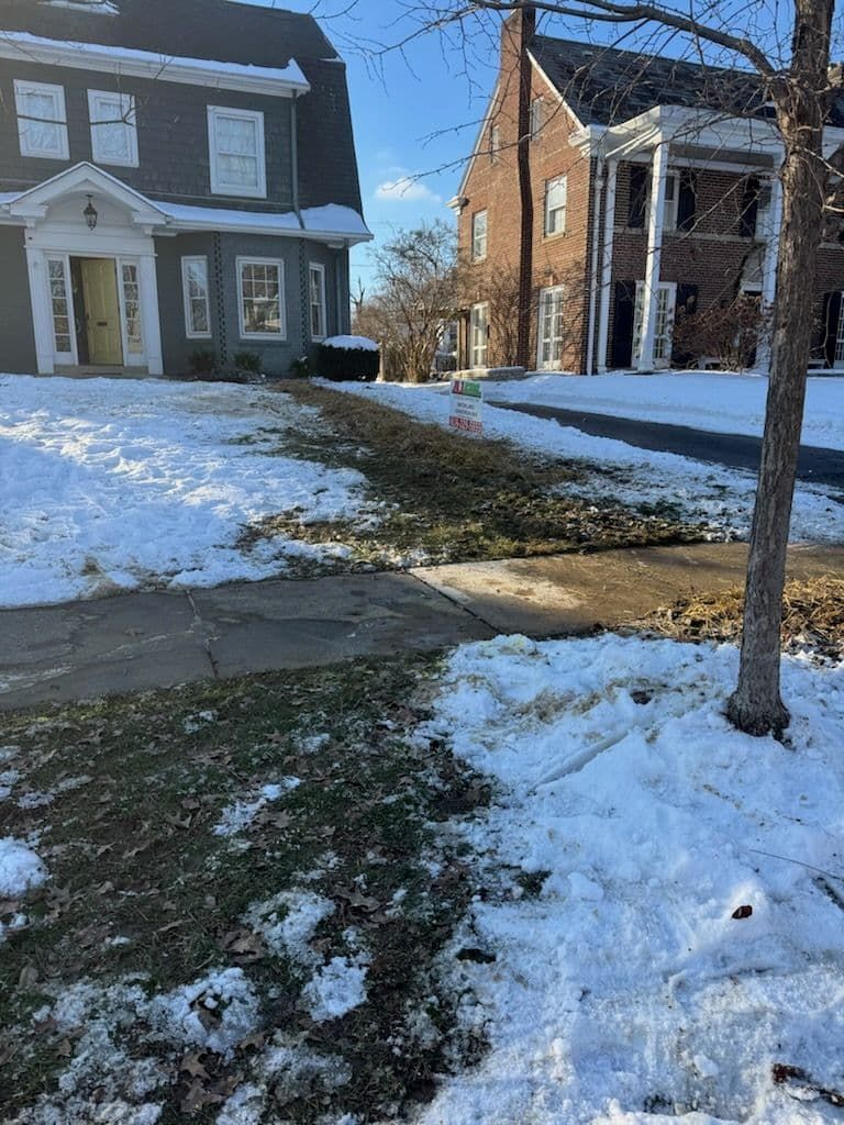 Snowy lawn and sidewalk in front of two-story homes. Patches of grass show through melting snow.