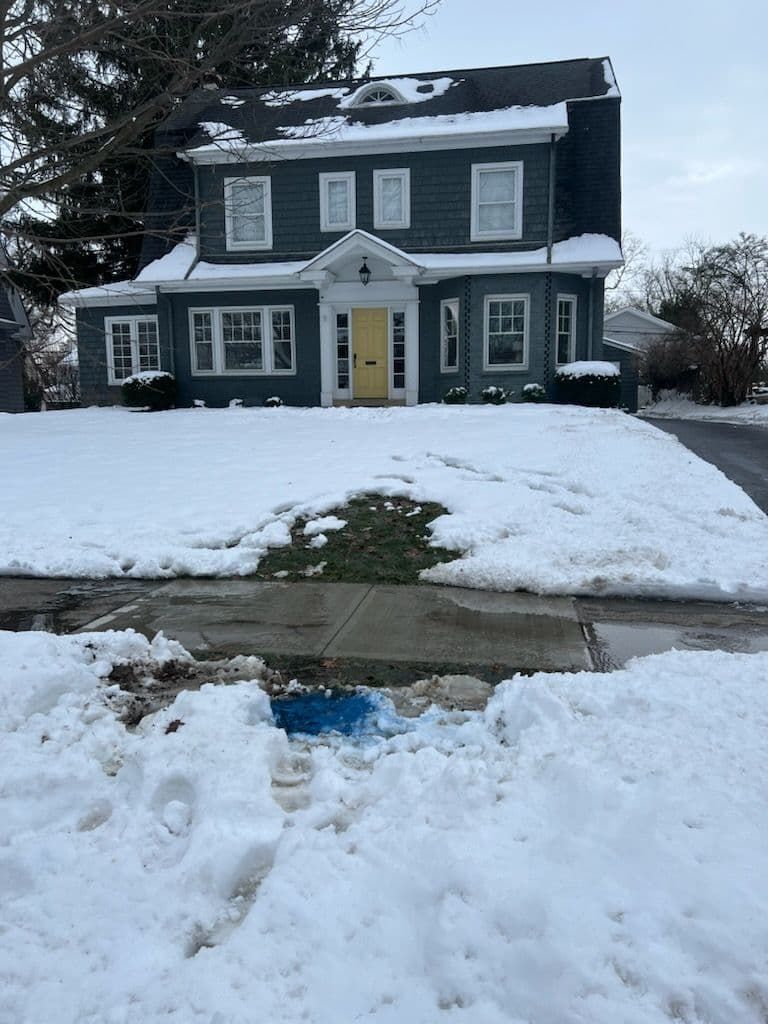 Snow-covered house with a yellow door, patches of grass visible, and snow-covered sidewalk.