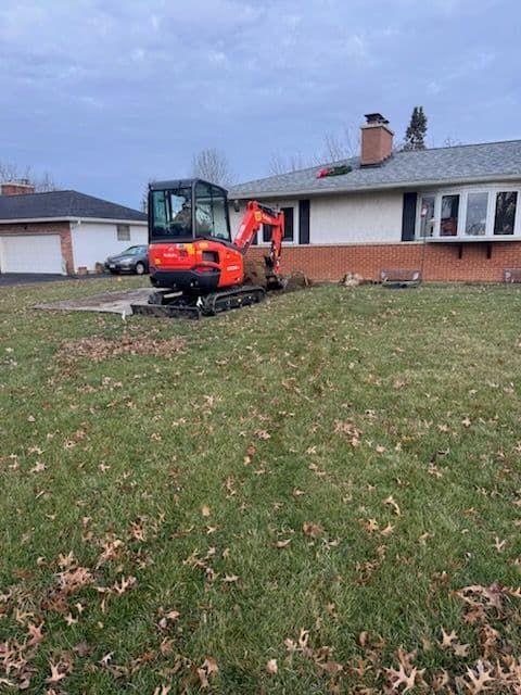 Orange excavator on grass near a brick house, digging. Overcast sky.
