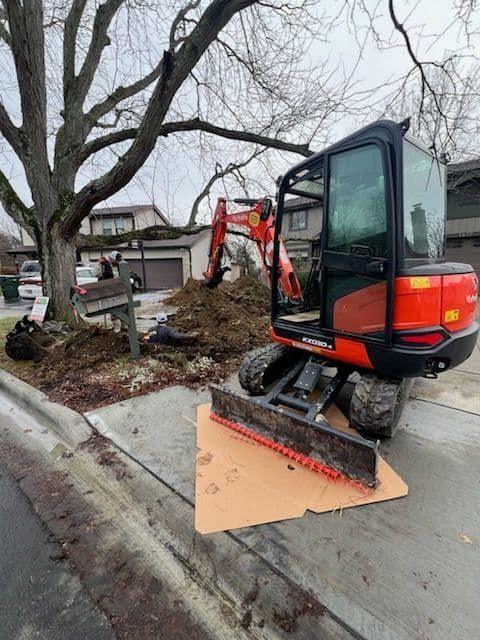 Orange excavator on a residential curb, digging near a tree, with a pile of dirt in the background.