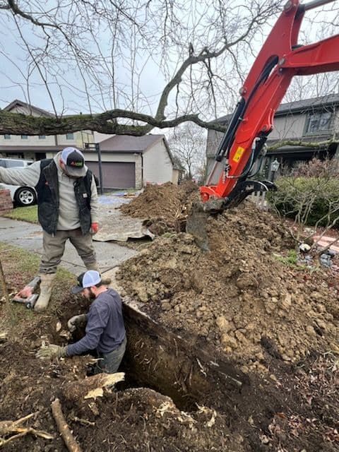 Two workers dig a trench with an excavator; one directs, one shovels. Outdoor setting.