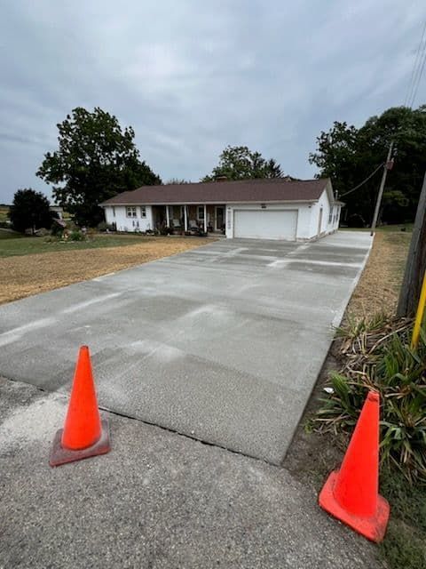 New concrete driveway leading to a white house with an attached garage, under a cloudy sky. Two orange safety cones are in the foreground.