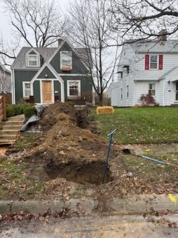 Dirt piled in front of a green house with a trench. A shovel stands upright. Another house is visible.