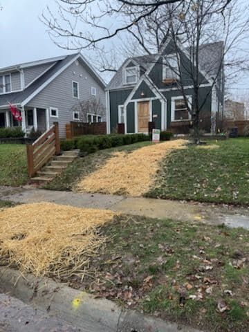 Residential street scene. A teal house with a straw-covered lawn. A gray house is to the left. Cloudy day.
