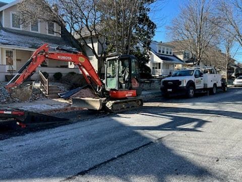 Mini excavator and work truck on a residential street. Orange excavator with arm extended; white truck parked alongside.