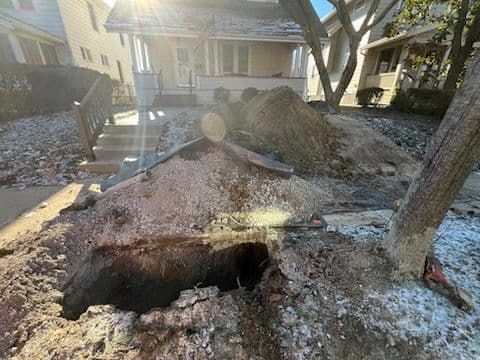 Large sinkhole in a front yard, near a house. Debris and a fallen tree are visible.