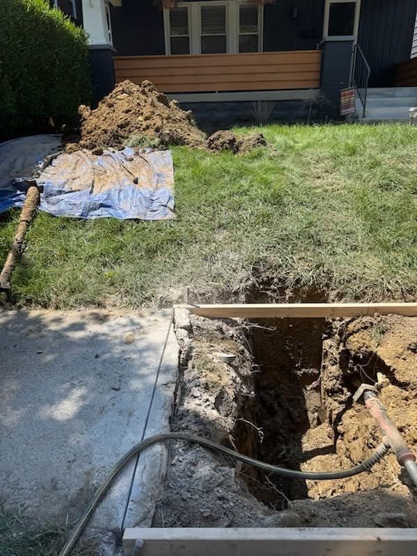 A trench dug in a grassy lawn near a sidewalk. Dirt pile and tarp nearby, house in background.