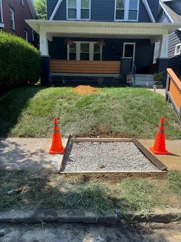 Sidewalk repair in progress; gravel-filled square, orange cones, house background.