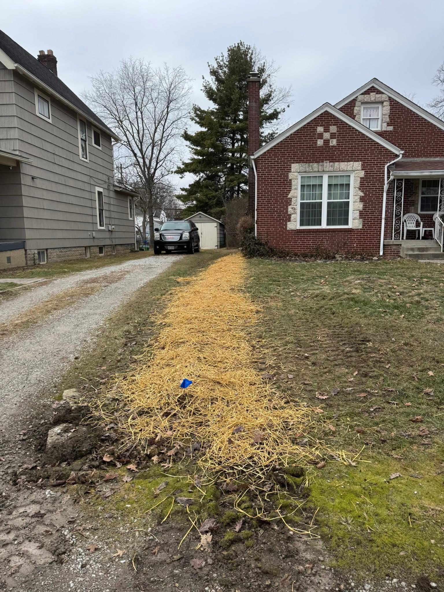 Gravel driveway with golden-colored debris alongside a house with red brick facade and a house with gray siding on a cloudy day.