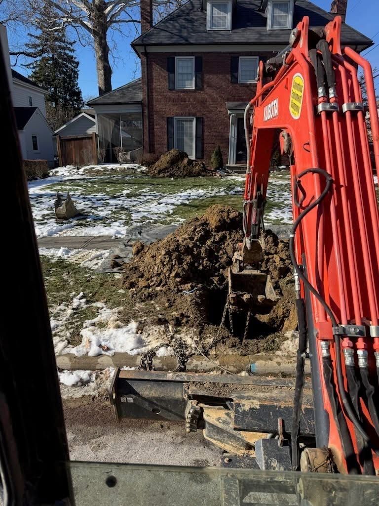 An excavator digging a hole in a front yard with some snow on the ground; a brick house in the background.
