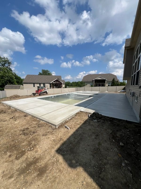 Construction of a pool and patio, with new concrete next to a partially built house, under a blue sky.