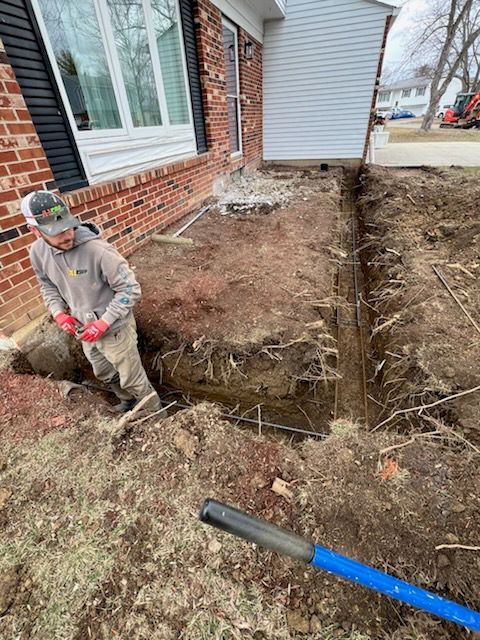Man digging a trench near a brick house. Brown soil, blue shovel, red gloves.