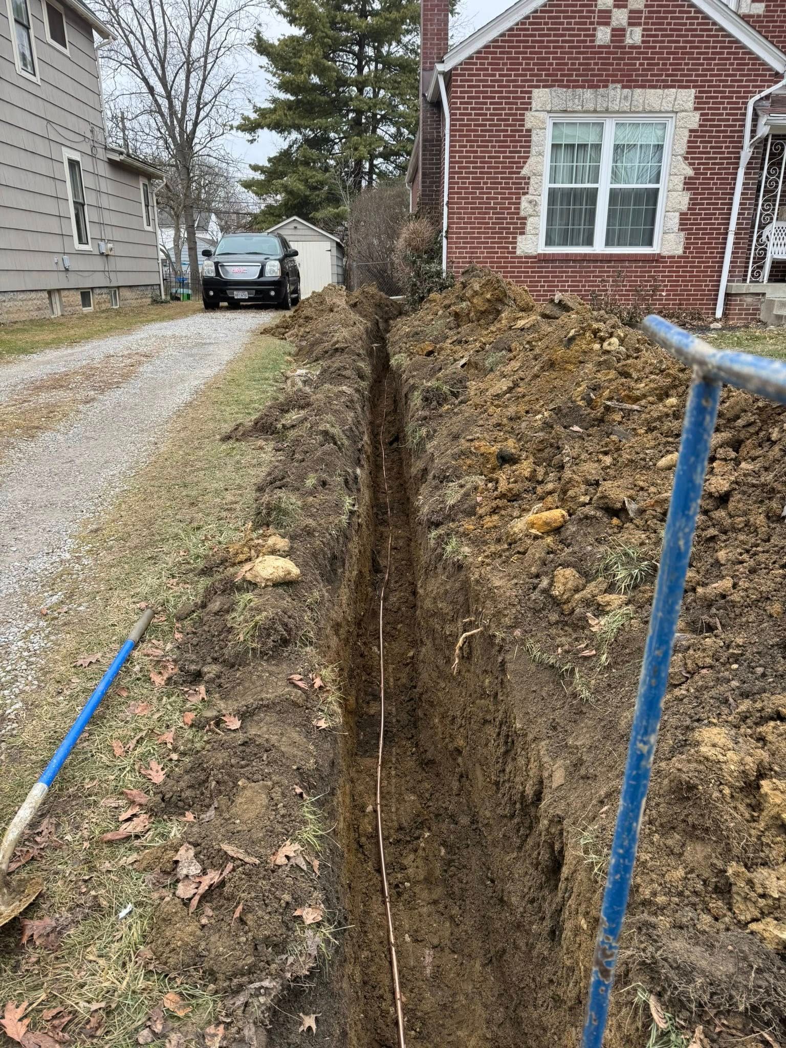 Trench dug in a yard next to a driveway and house; blue handled tools are visible.