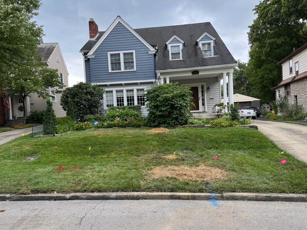 Blue house with white porch and dormers, on a grassy lawn with bushes and flowers.