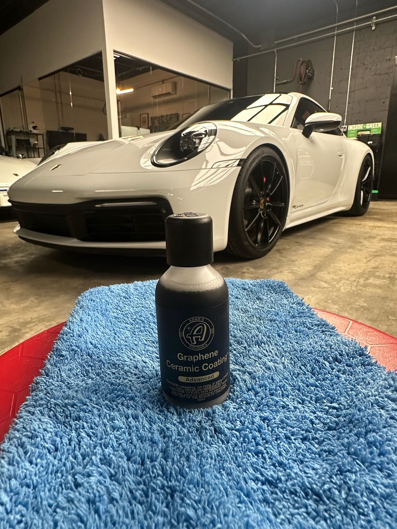 A bottle of detailing product sits on a blue microfiber cloth in front of a white Porsche in a garage.