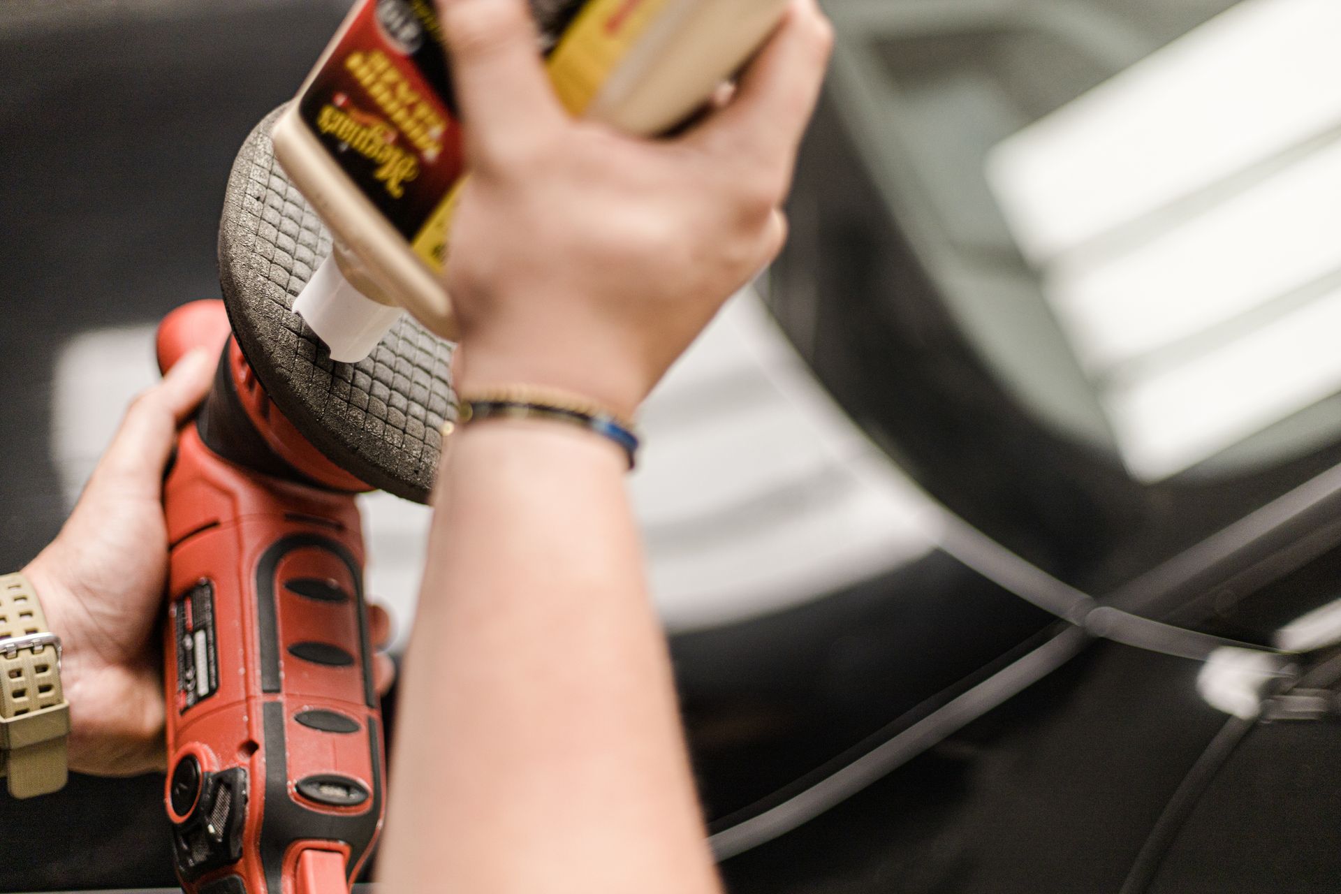 A person applies polishing compound from a bottle onto the pad of a red electric buffer.