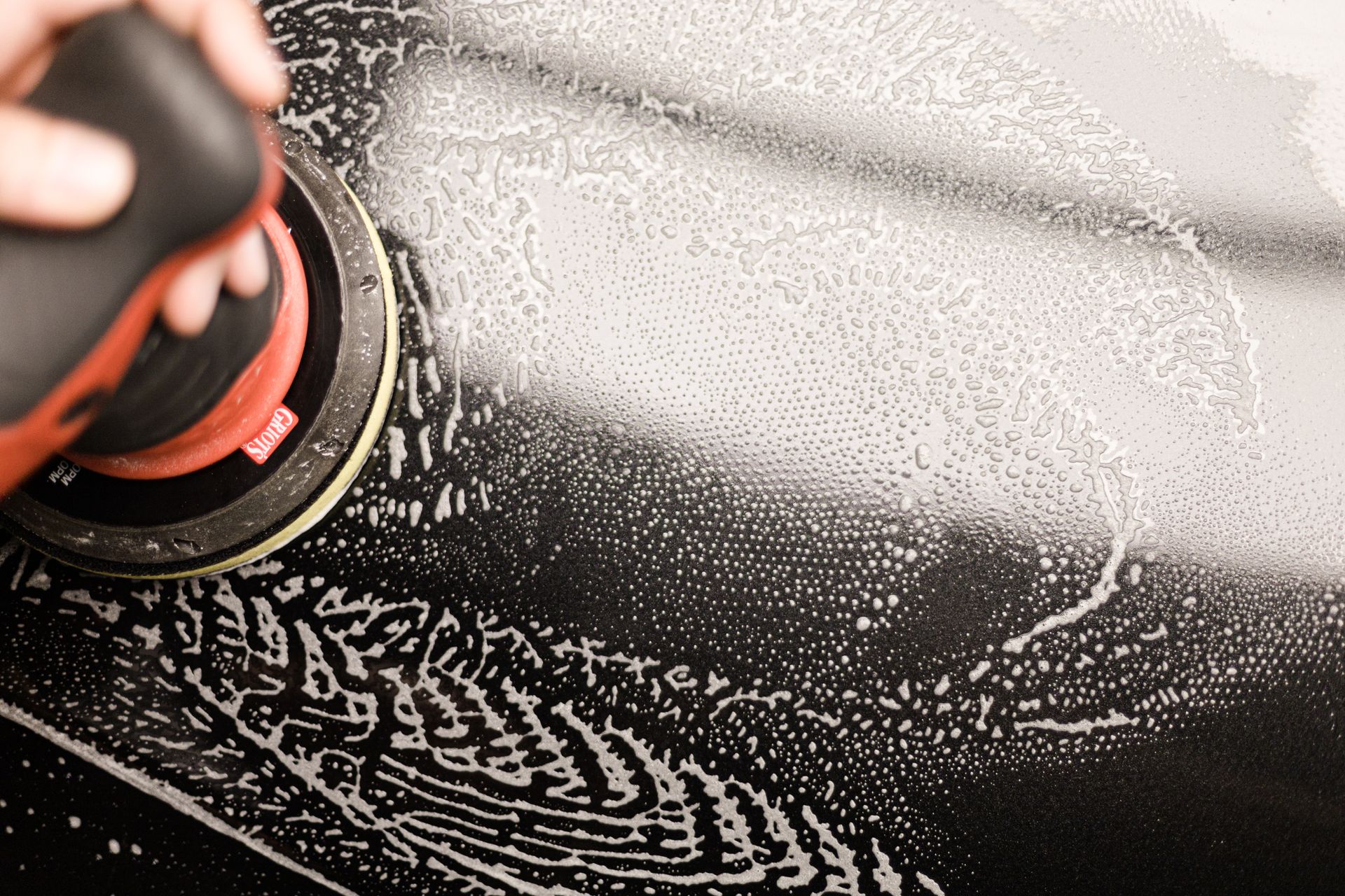 A hand uses a motorized power buffer to polish a dark, sudsy car surface.