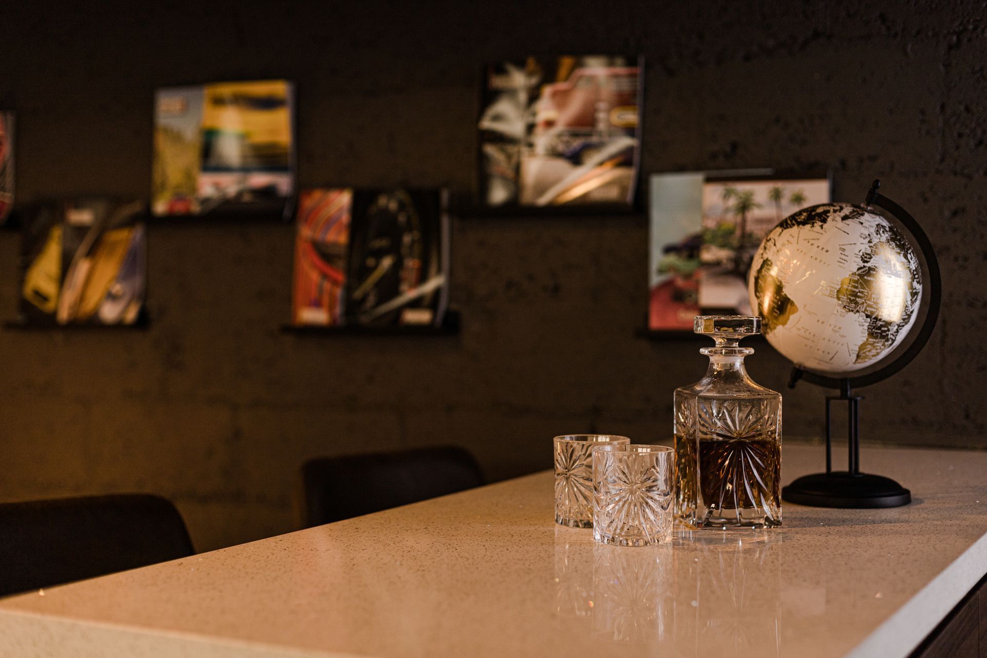 A white countertop holds two empty crystal glasses, a decanter of amber liquid, and a small globe against a dark wall.