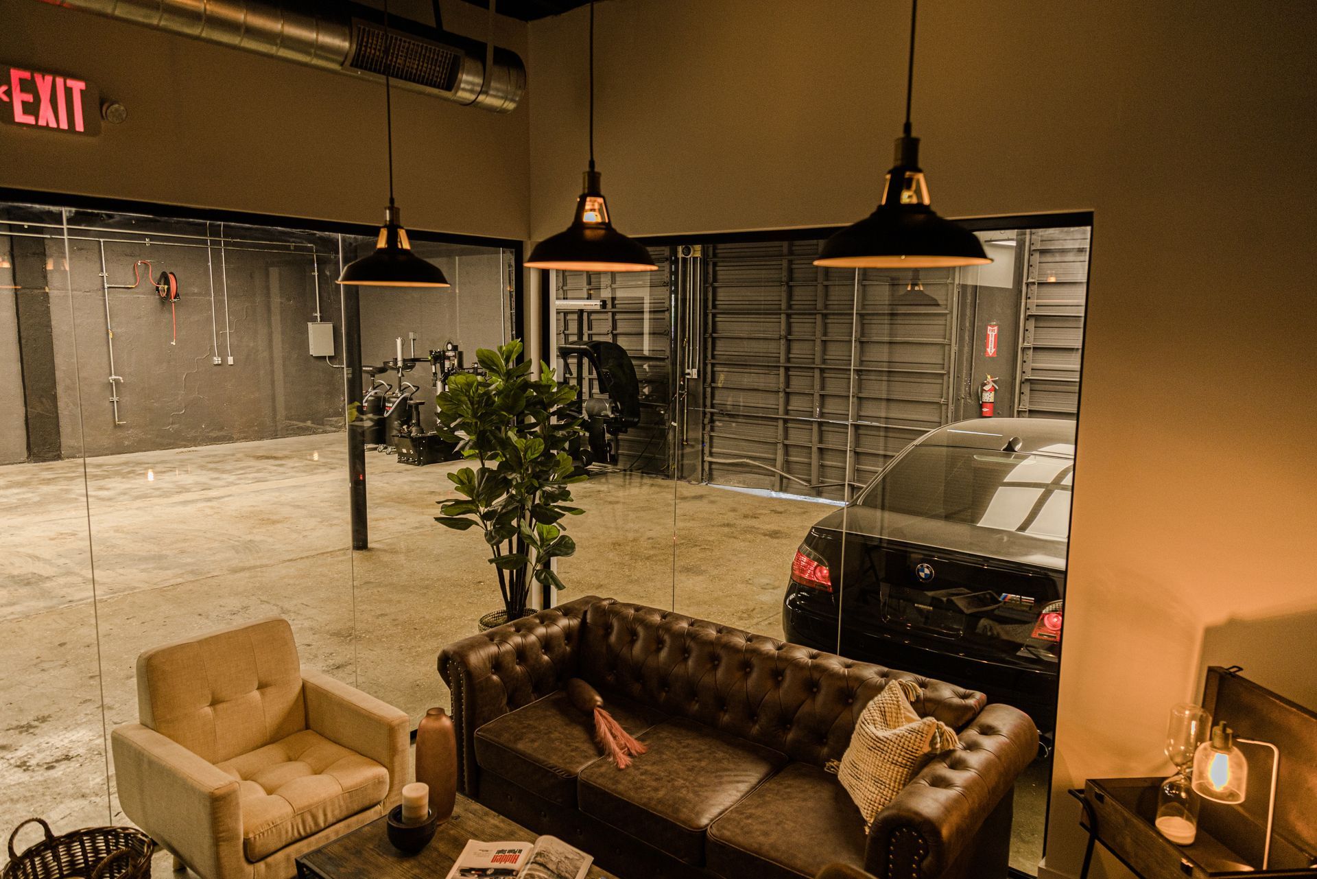 Modern industrial lounge area with a brown leather sofa, beige chair, potted plant, and a car visible through a glass wall.