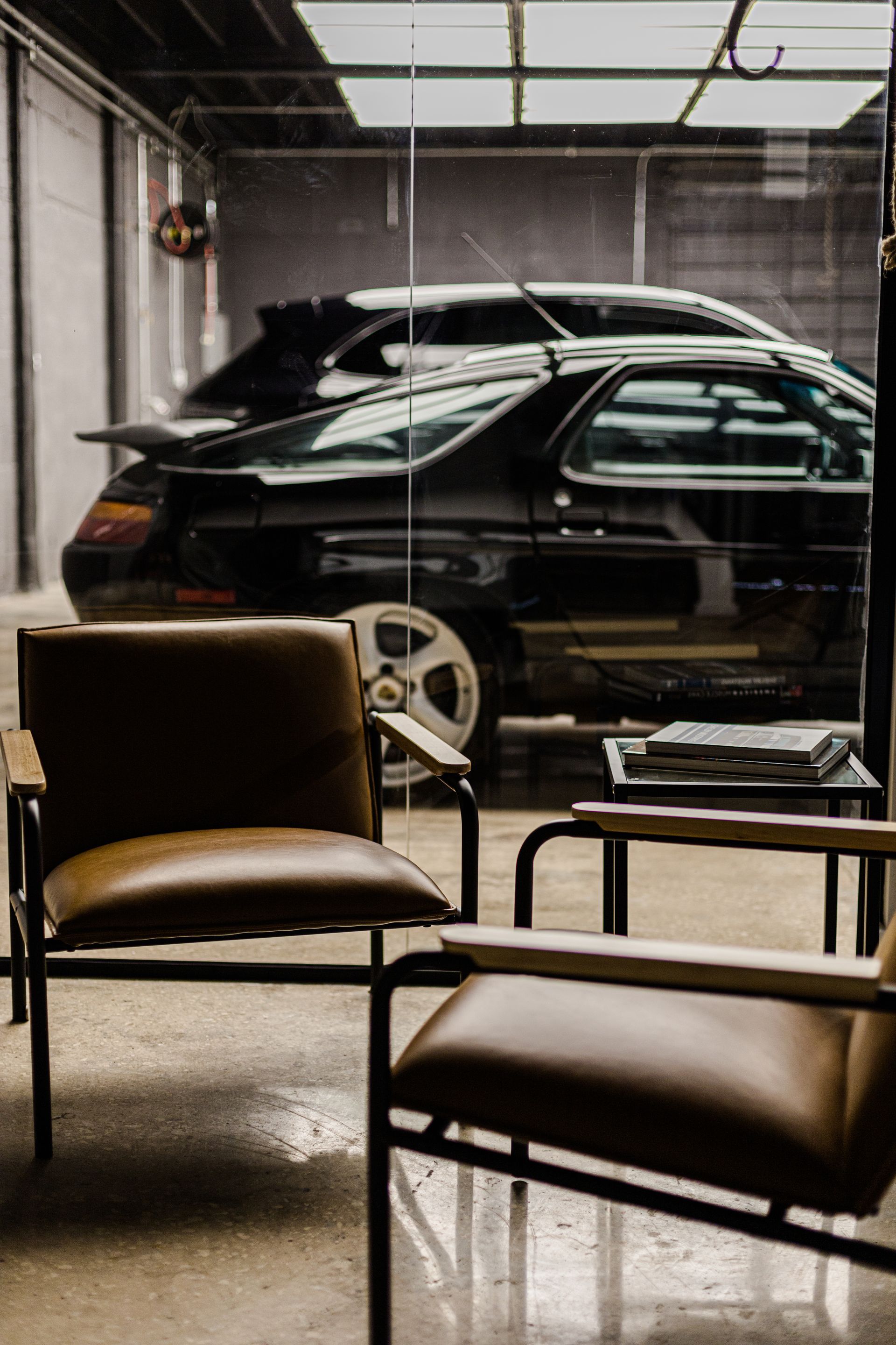 Two leather armchairs in a modern lounge, with a view of a black sports car parked in a garage through a glass wall.