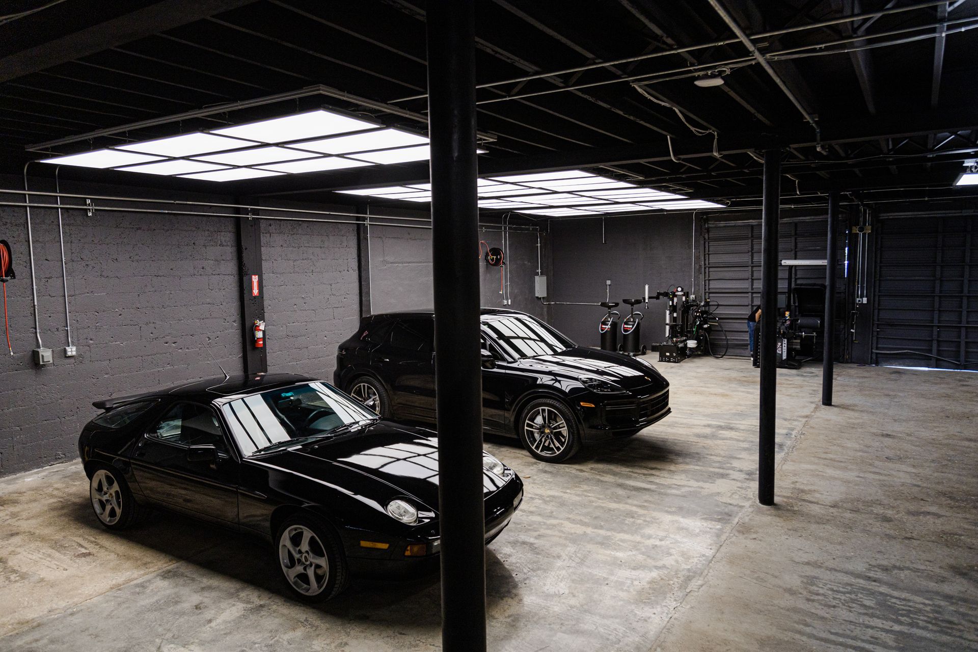 Two black sports cars parked in a dimly lit industrial-style garage with bright overhead grid lighting.