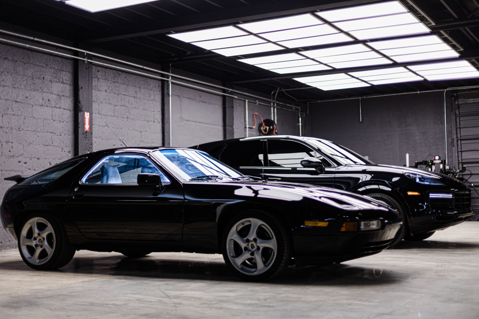 A black Porsche 928 sports car parked in front of a black SUV in a garage with a paneled skylight and concrete walls.