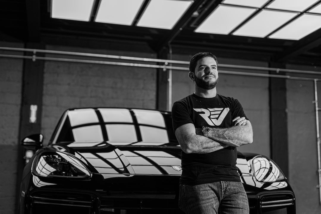 Man with arms crossed standing in front of a black car in a garage.