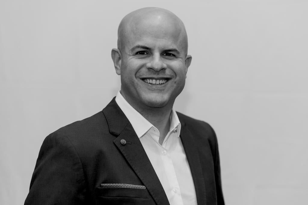 Man with a shaved head smiles, wearing a suit jacket and white shirt against a white wall.