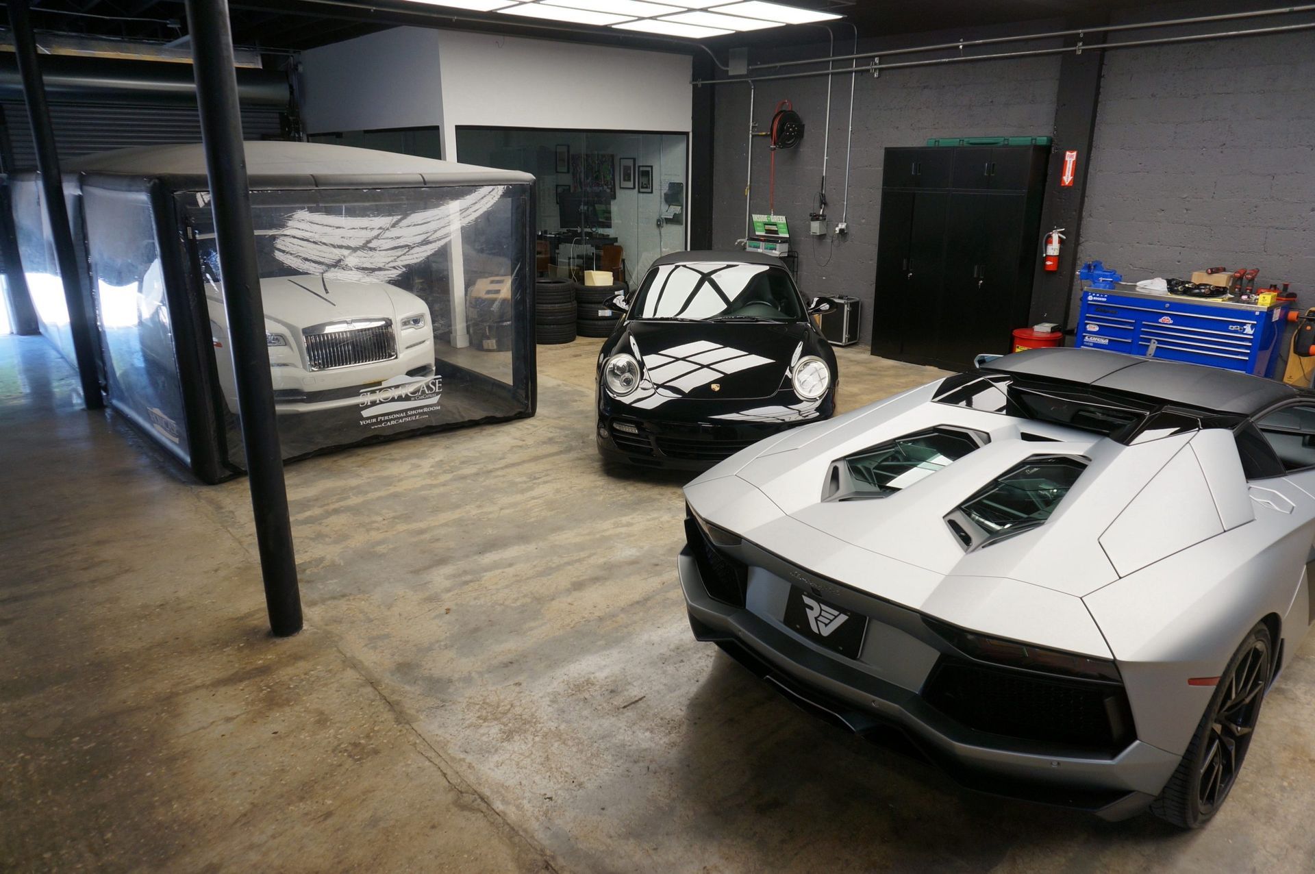 Cars in a garage: a silver Lamborghini, a black Smart car, and a white car in a clear enclosure.