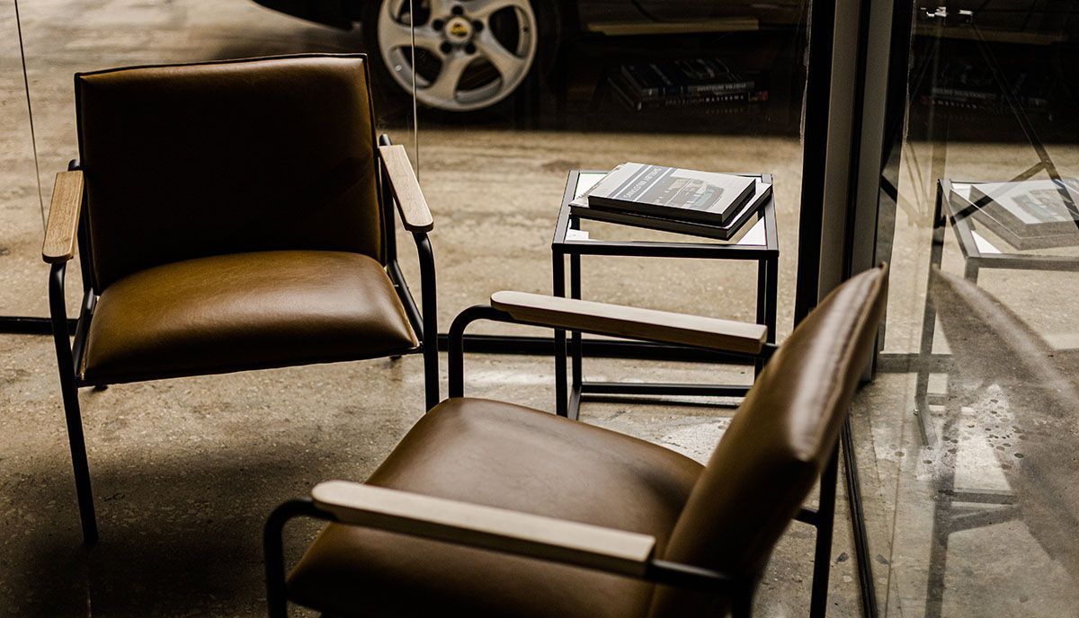 Two brown leather armchairs sit near a small table with reading material, inside a room; car wheel visible through window.